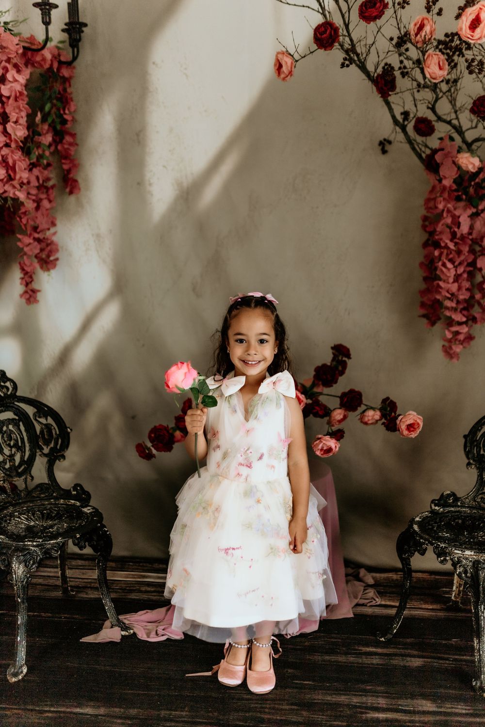 Young girl in floral white dress holds pink rose, posing before vintage chairs and flower backdrop.