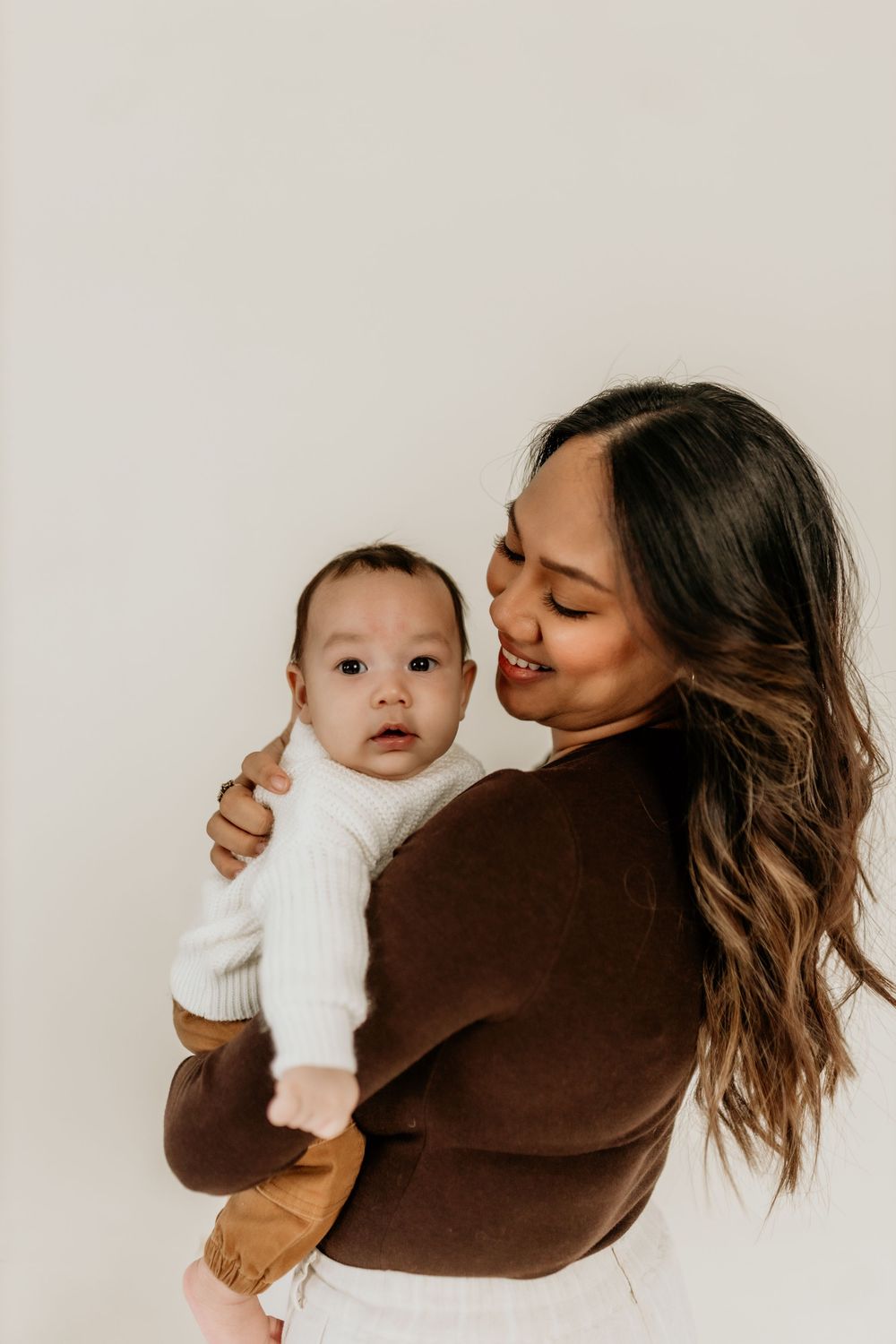 A tender series of photos showing a parent in brown sweater holding and embracing a baby in white clothing against white backdrop.