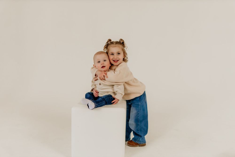 Two young siblings pose together on a white cube, older toddler girl hugging baby brother in a bright studio.