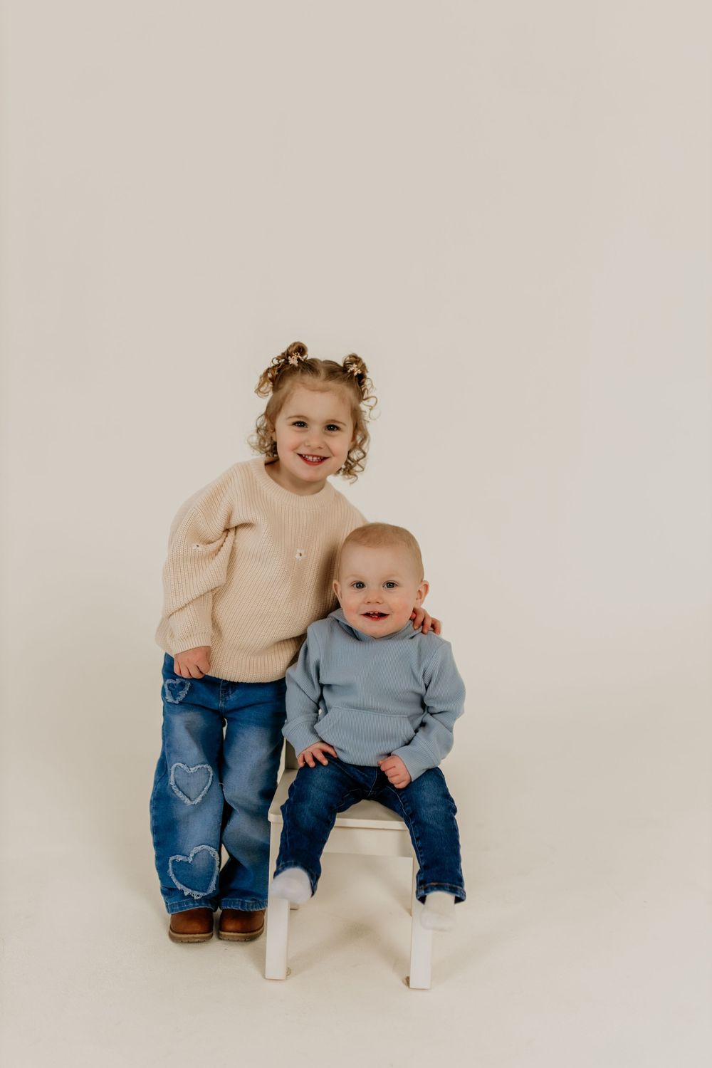 Two young siblings pose together, older girl in beige sweater and heart jeans stands behind baby boy in blue top on white stool.