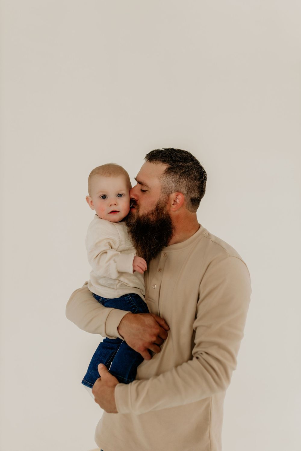 Bearded father in beige sweater kissing his baby on the cheek against a white background.
