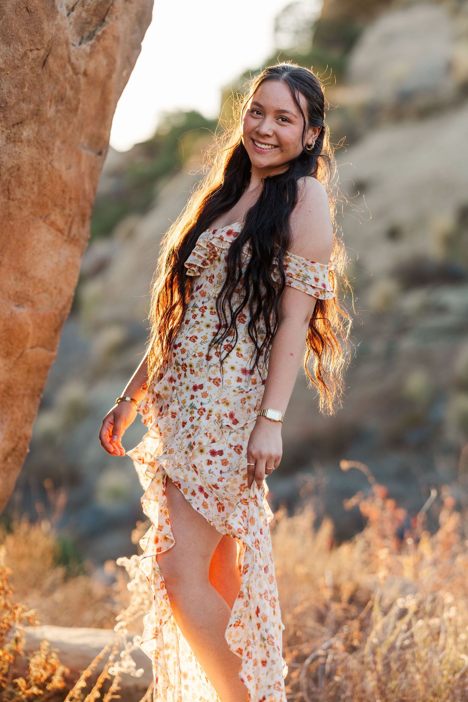 A person in a floral dress poses playfully in golden sunlight among tall grass during a sunset photoshoot.