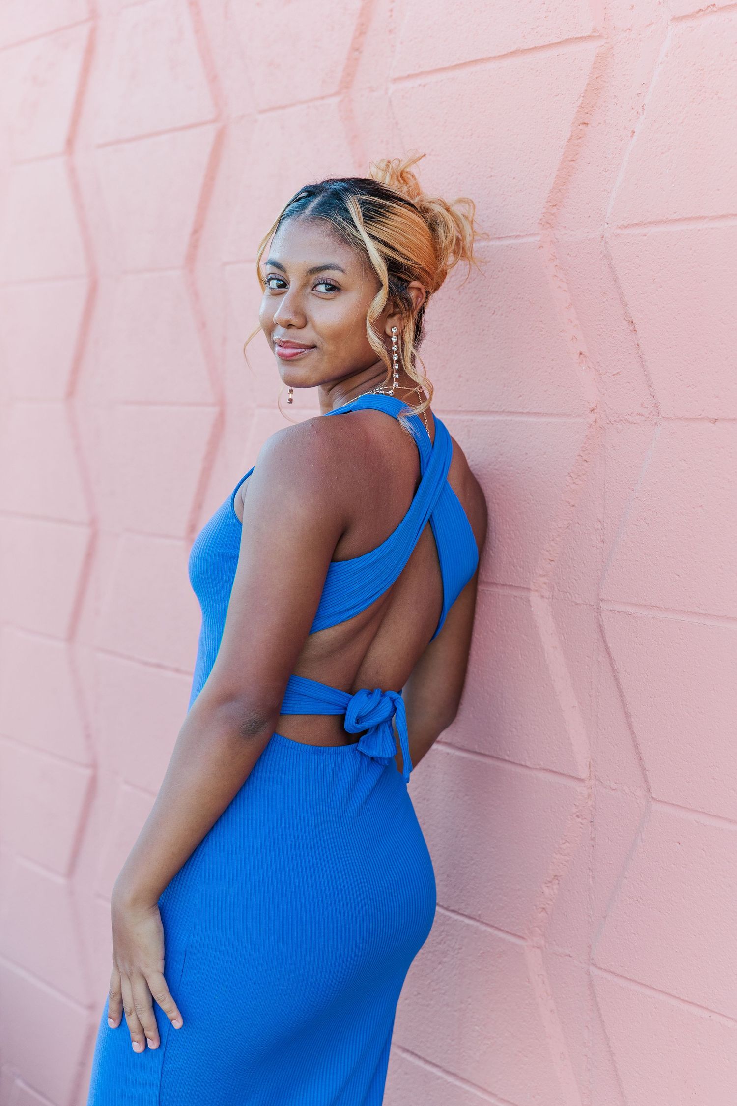 A woman in a bright blue fitted dress poses against a pink wall showing different angles of the garment.