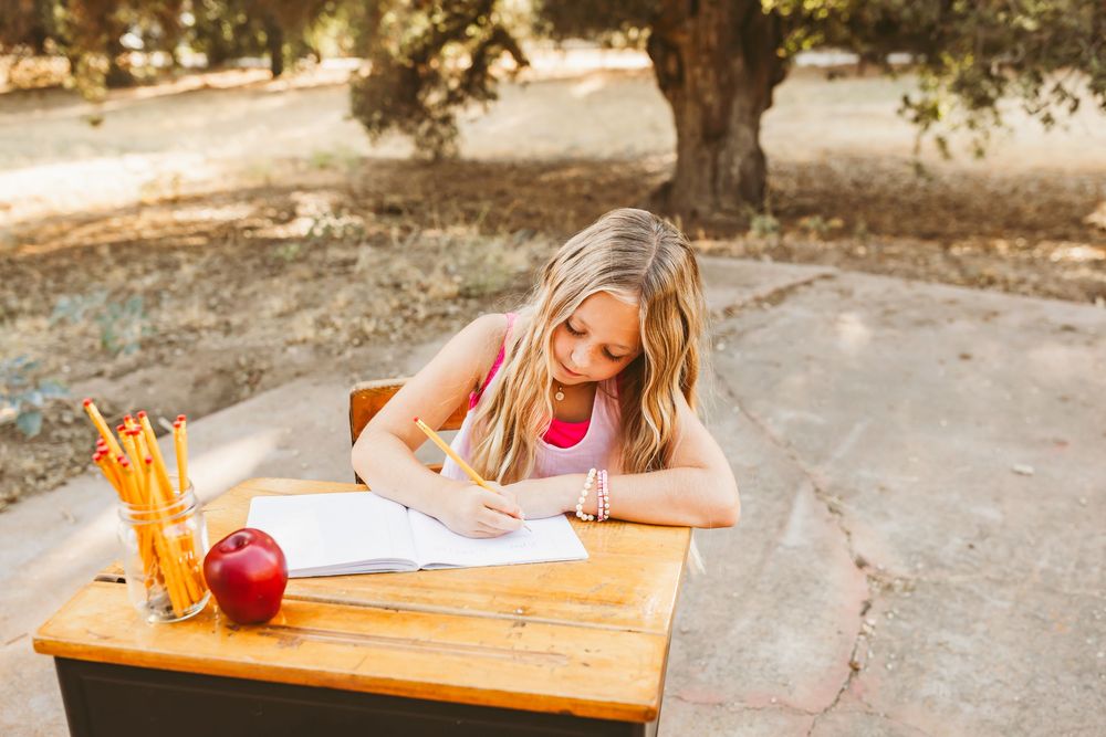 Student writing in notebook at wooden desk outdoors with red apple and orange drink in sunlit setting.
