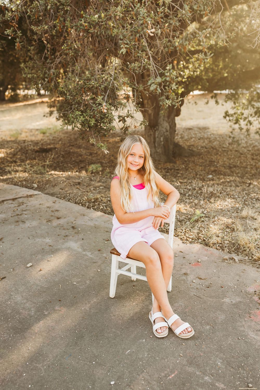 A young student sits on a white chair outdoors wearing a light pink dress and white sandals in a sunlit garden setting.