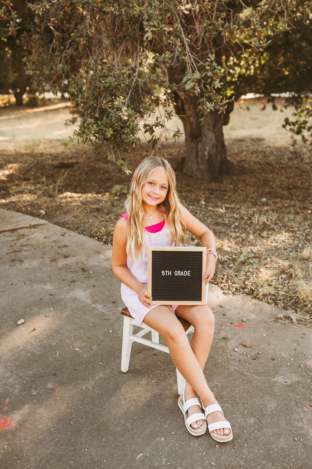 A student poses with a chalkboard sign while sitting on a white chair under an olive tree.