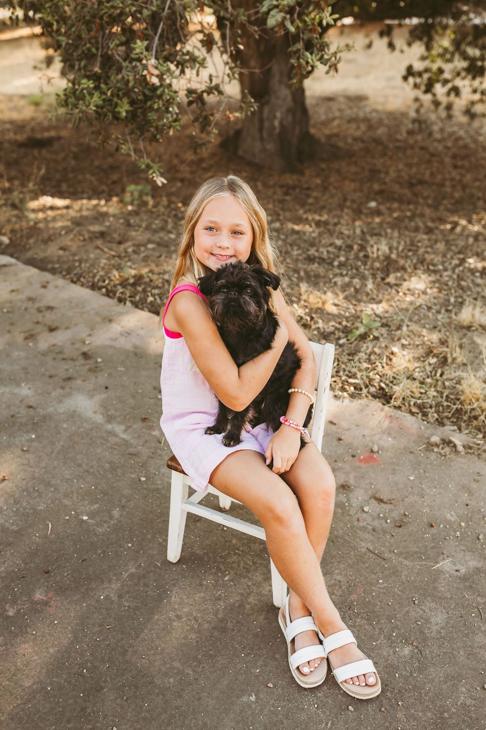 A person wearing a pink dress and white sandals cuddles with a black dog while sitting on a chair outside.