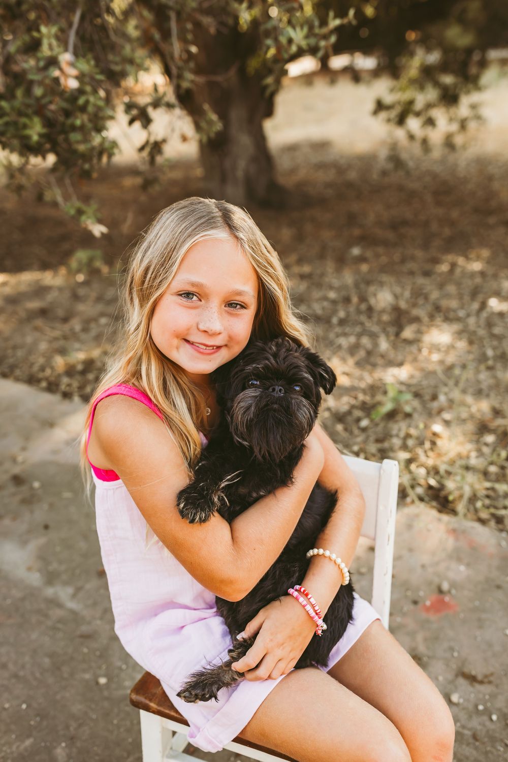 A young person in a pink dress sits on a white chair while holding a small black dog outdoors.