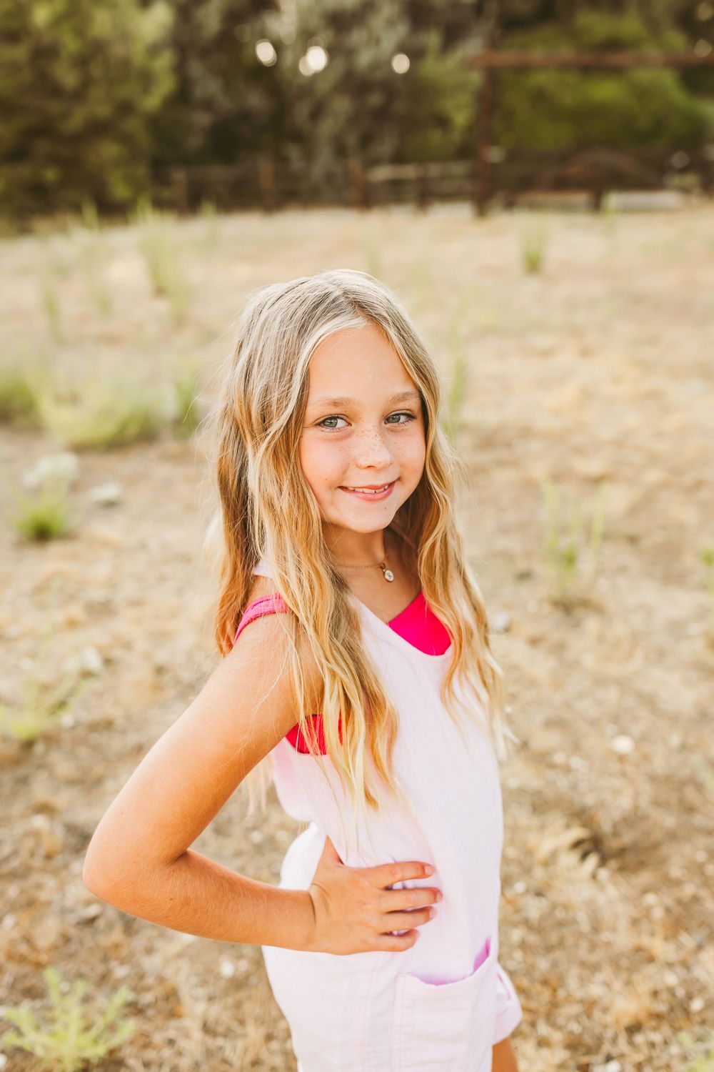 A young child wearing a white and pink sleeveless top poses playfully in an outdoor setting with dry grass and trees.