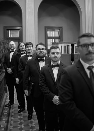 Groomsmen in black tuxedos walk through an elegant hallway with arched ceilings.
