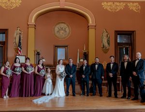 Wide panoramic shot of a wedding party lined up in a grand ballroom with ornate decor.