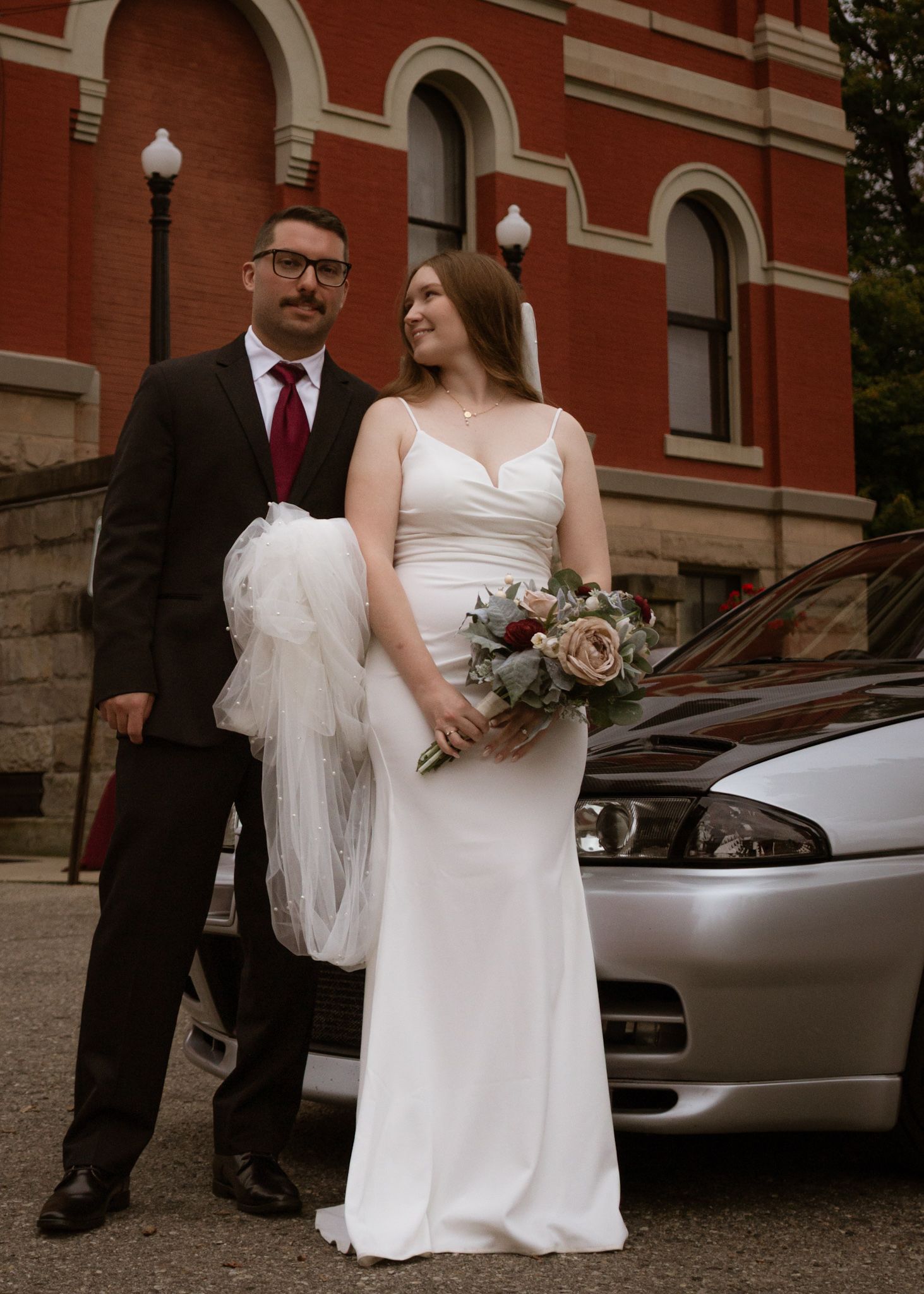 Wedding couple poses outside a historic red brick building with arched windows.