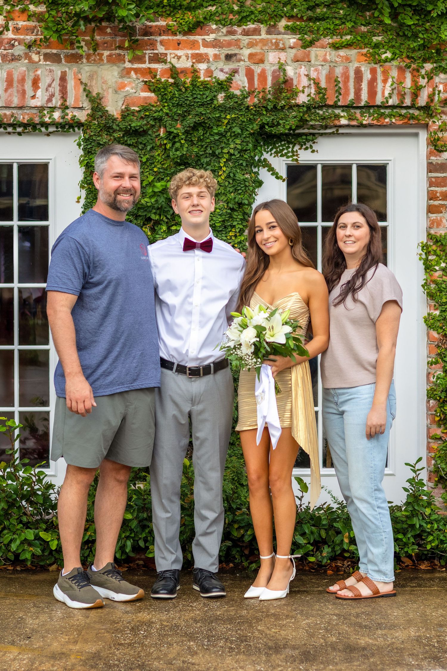 Family group poses together outdoors against brick wall with green ivy in casual summer attire.
