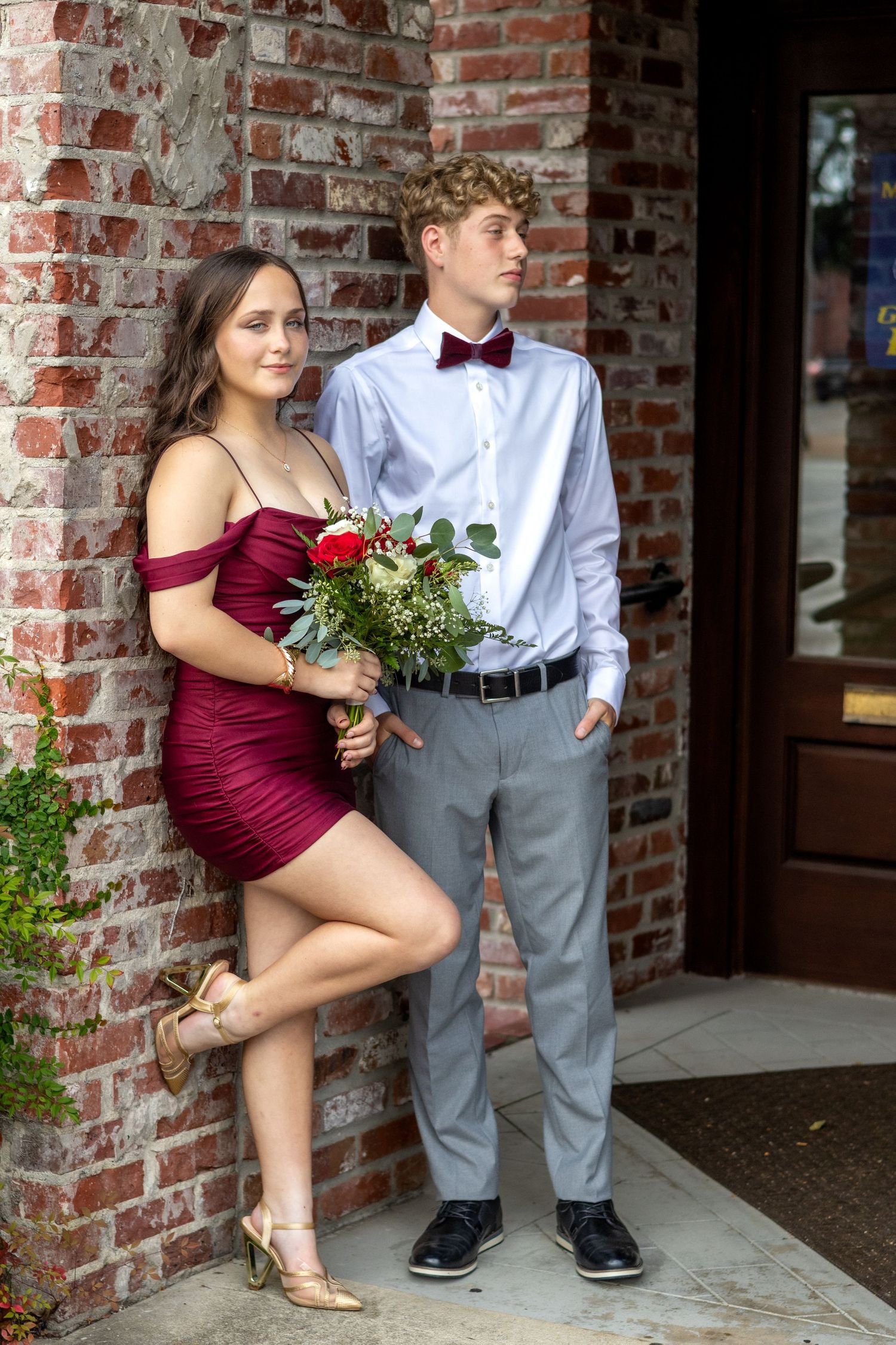 A couple in formal attire poses for photos against a brick wall, with her in burgundy and him in gray slacks.