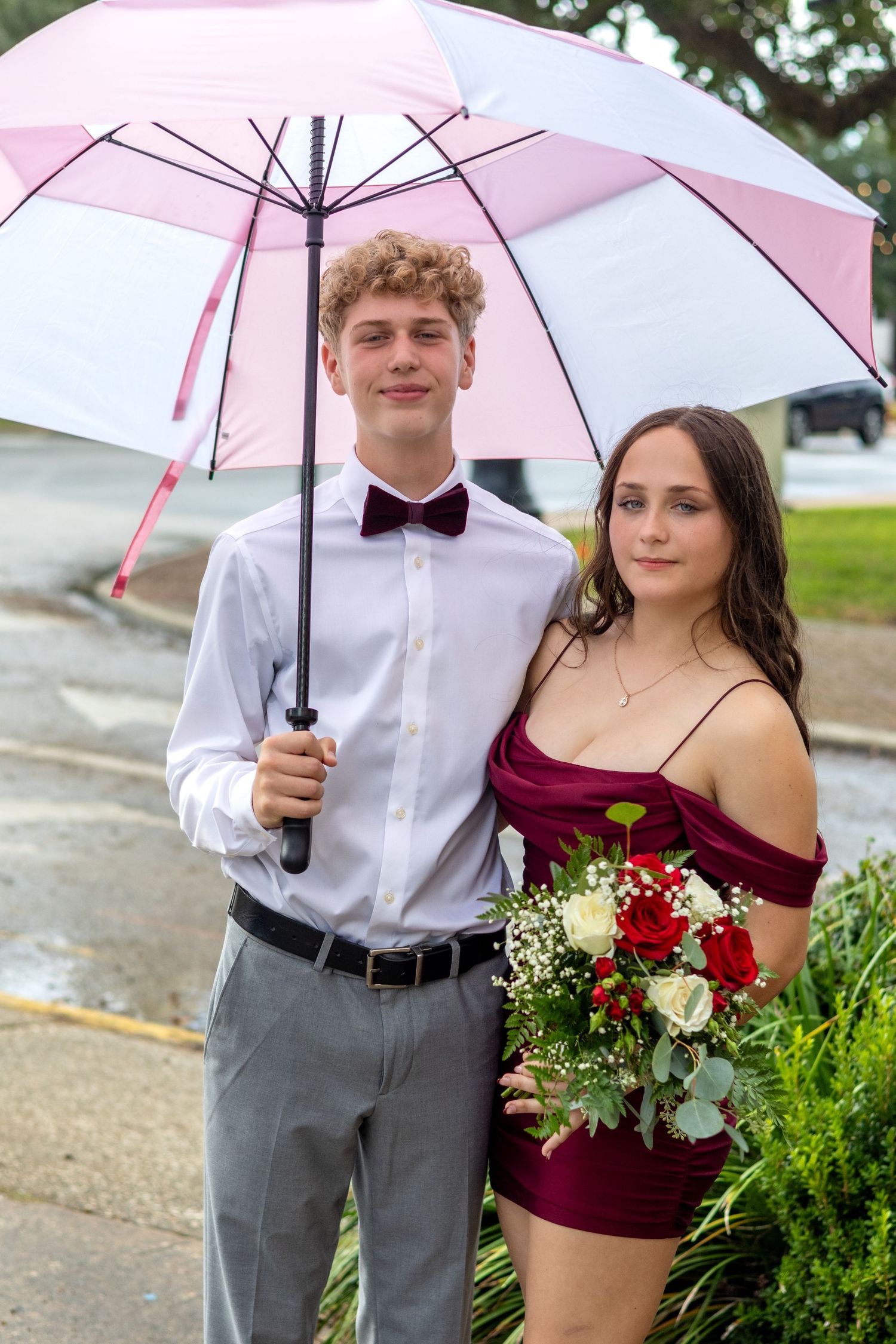 A couple stands under a pink umbrella in formal attire with burgundy accents and floral arrangements.