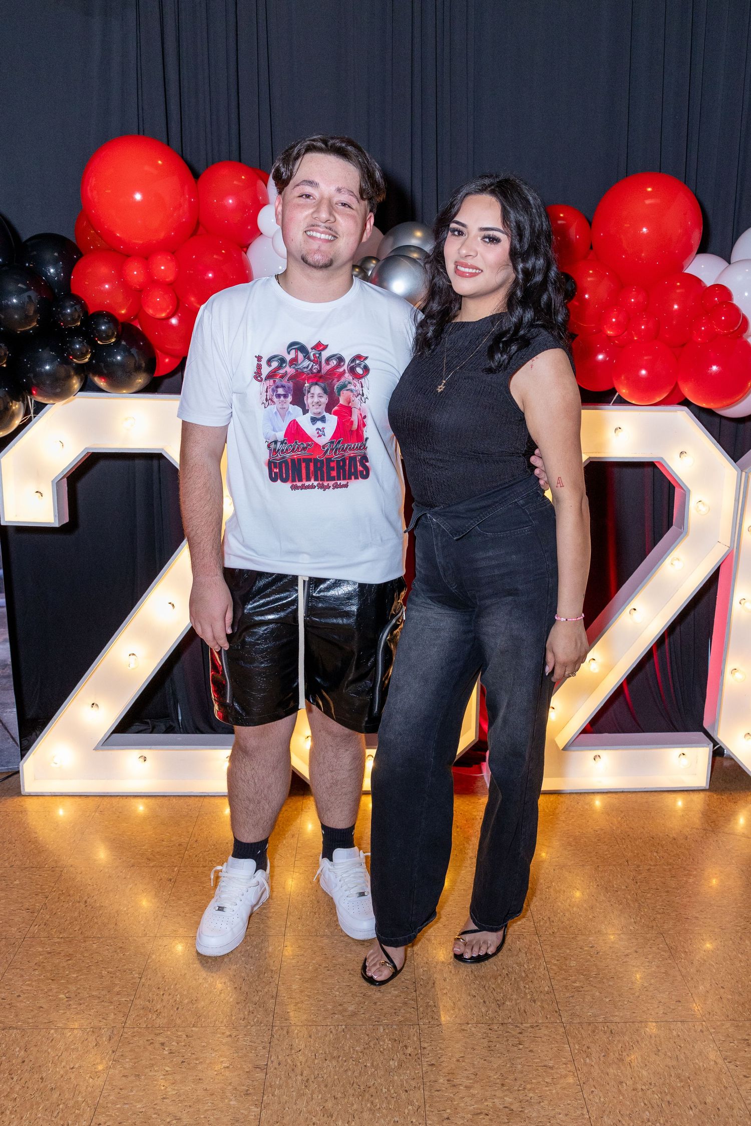 Two people posing together at a party with red, black, and white balloon decorations and illuminated number displays.