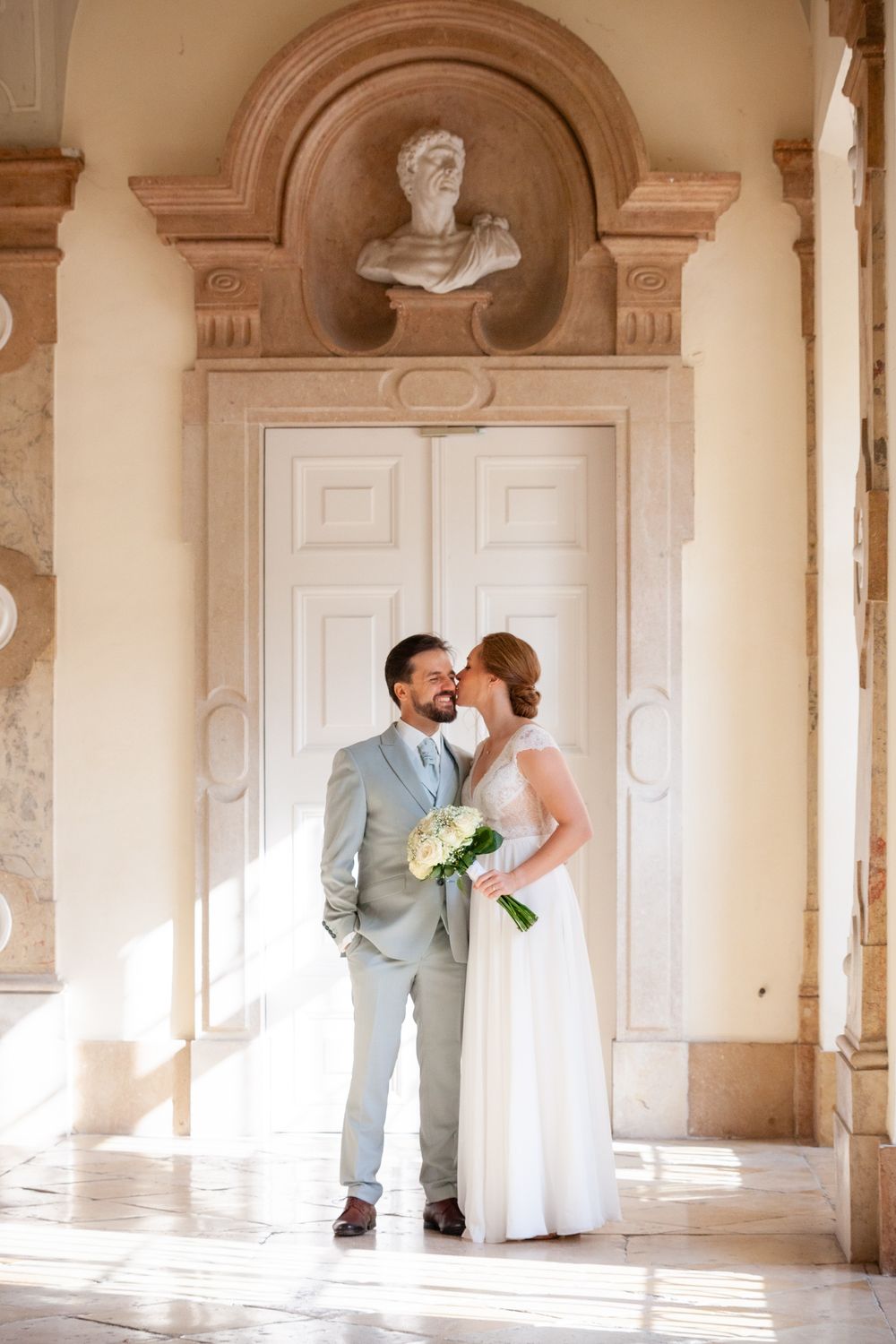 A romantic wedding couple embracing in an ornate colonnade with arched doorways and cream colored walls.
