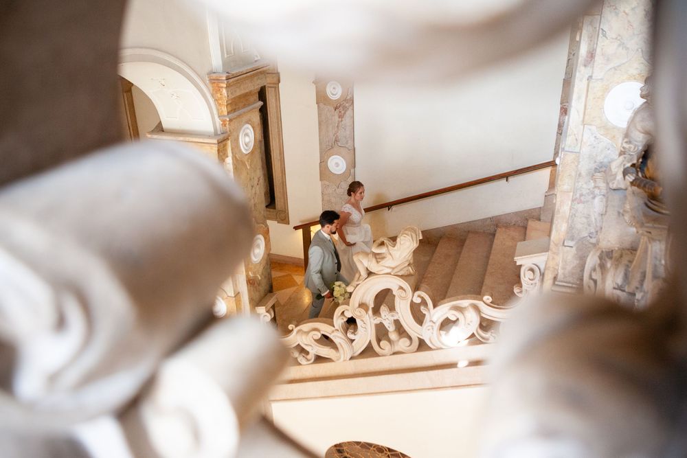 A person signs documents at an ornate interior staircase with decorative architectural details.