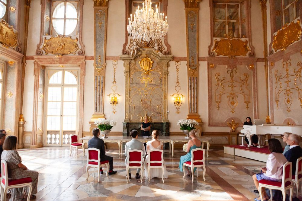 Ornate baroque ballroom with crystal chandeliers, gilded walls, and red velvet chairs.