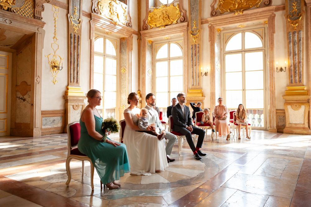 Interior of an opulent baroque palace hall with golden details, arched windows, and people exploring the space.