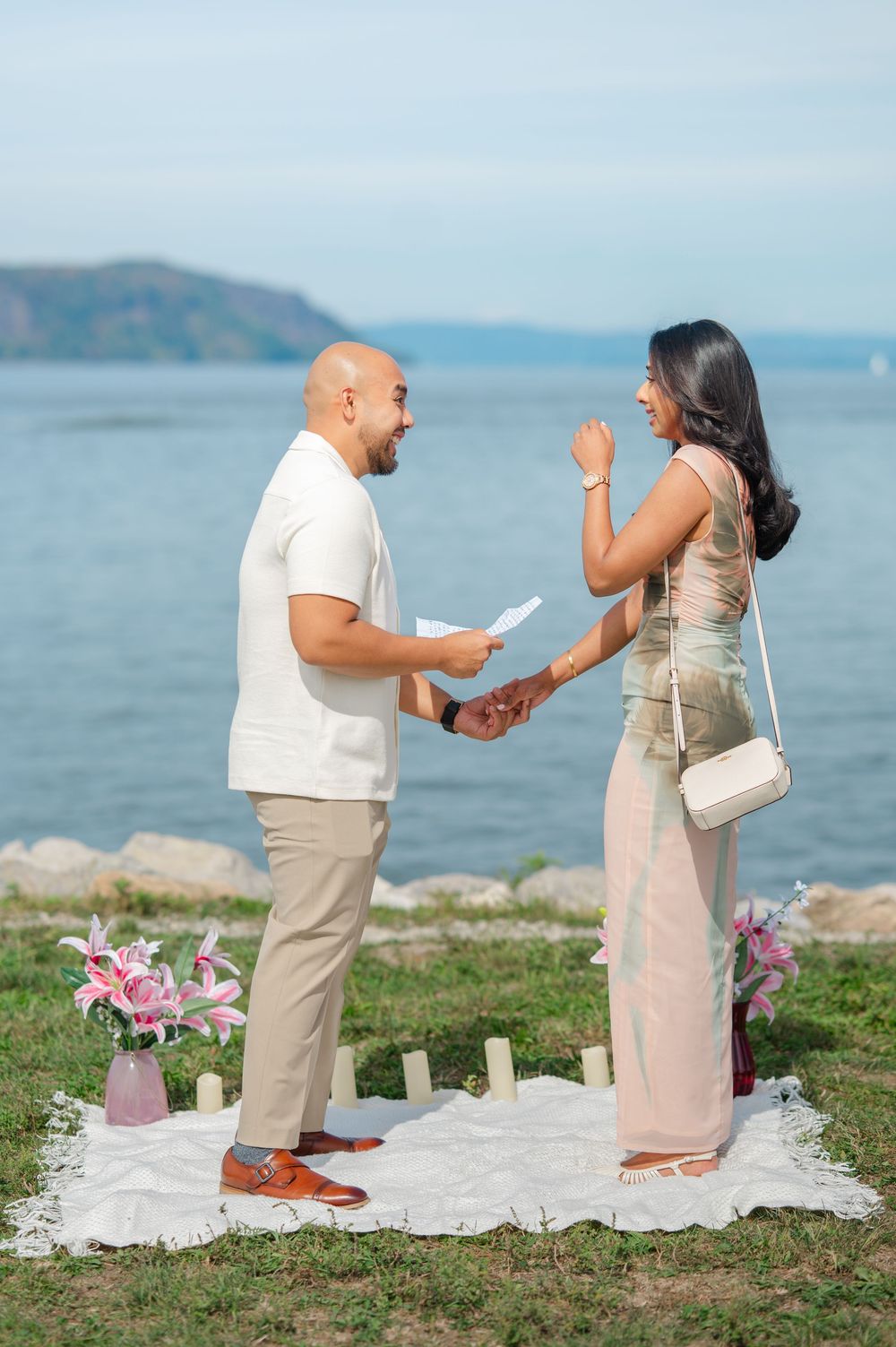 A couple shares an emotional surprise proposal moment next to the Hudson River with a white blanket and flowers.