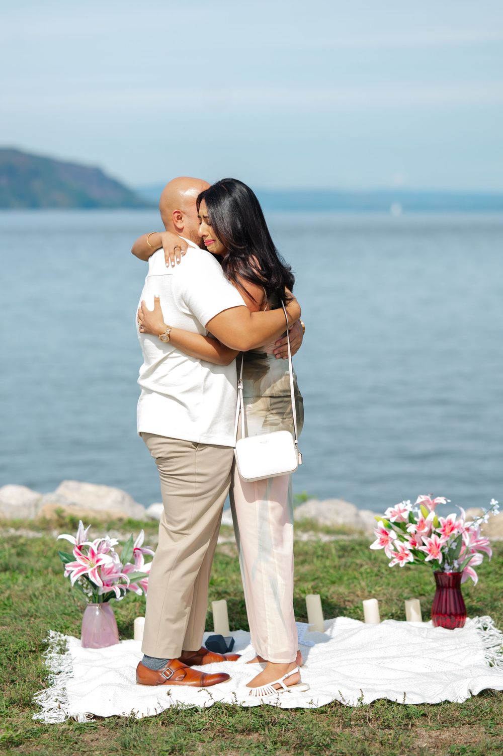 A couple hugs during a romantic riverside proposal with flowers and decor set up on a grassy area overlooking the Hudson River.