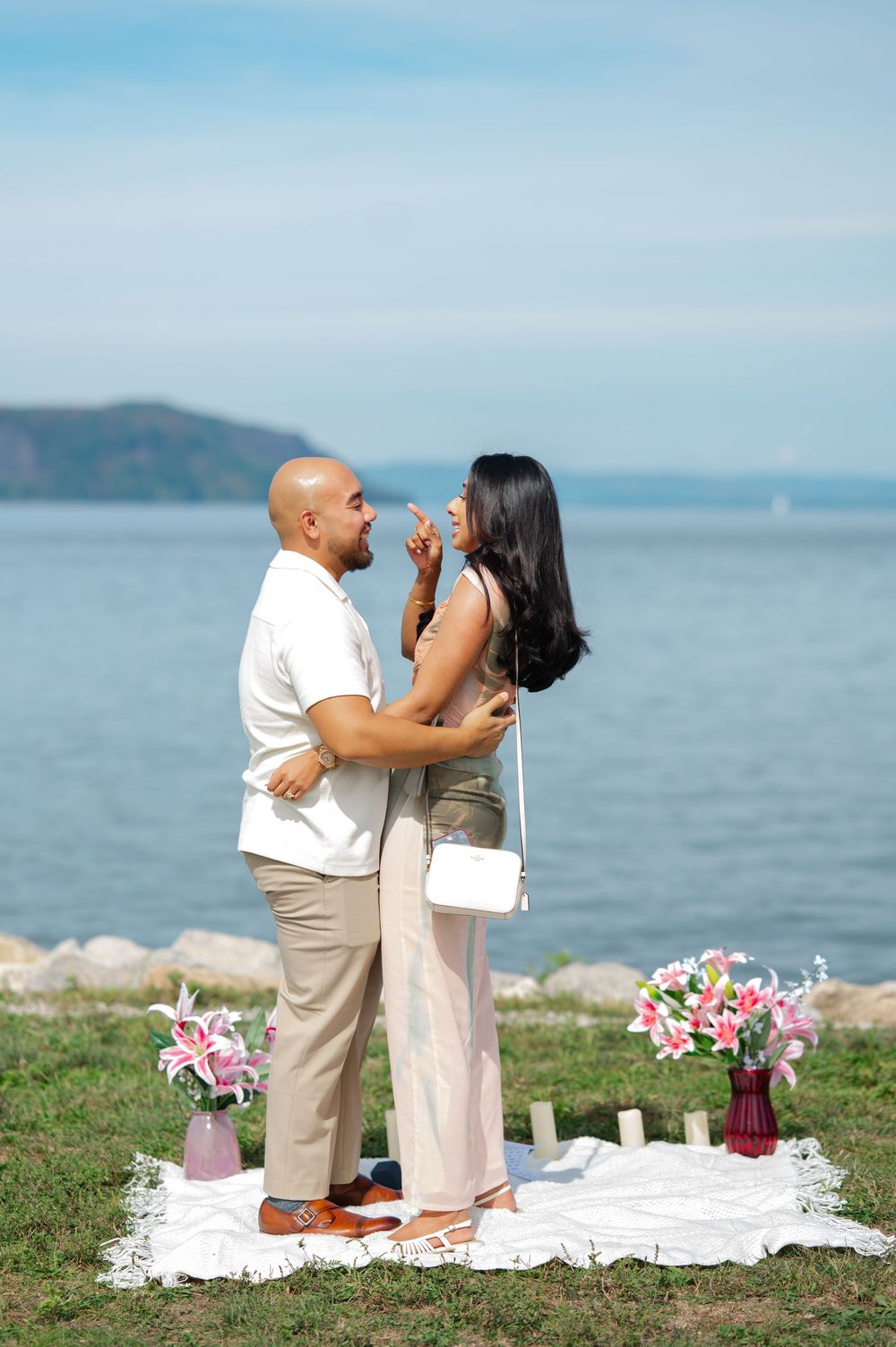 A romantic lakeside proposal scene with mountains in the background and pink floral decorations.