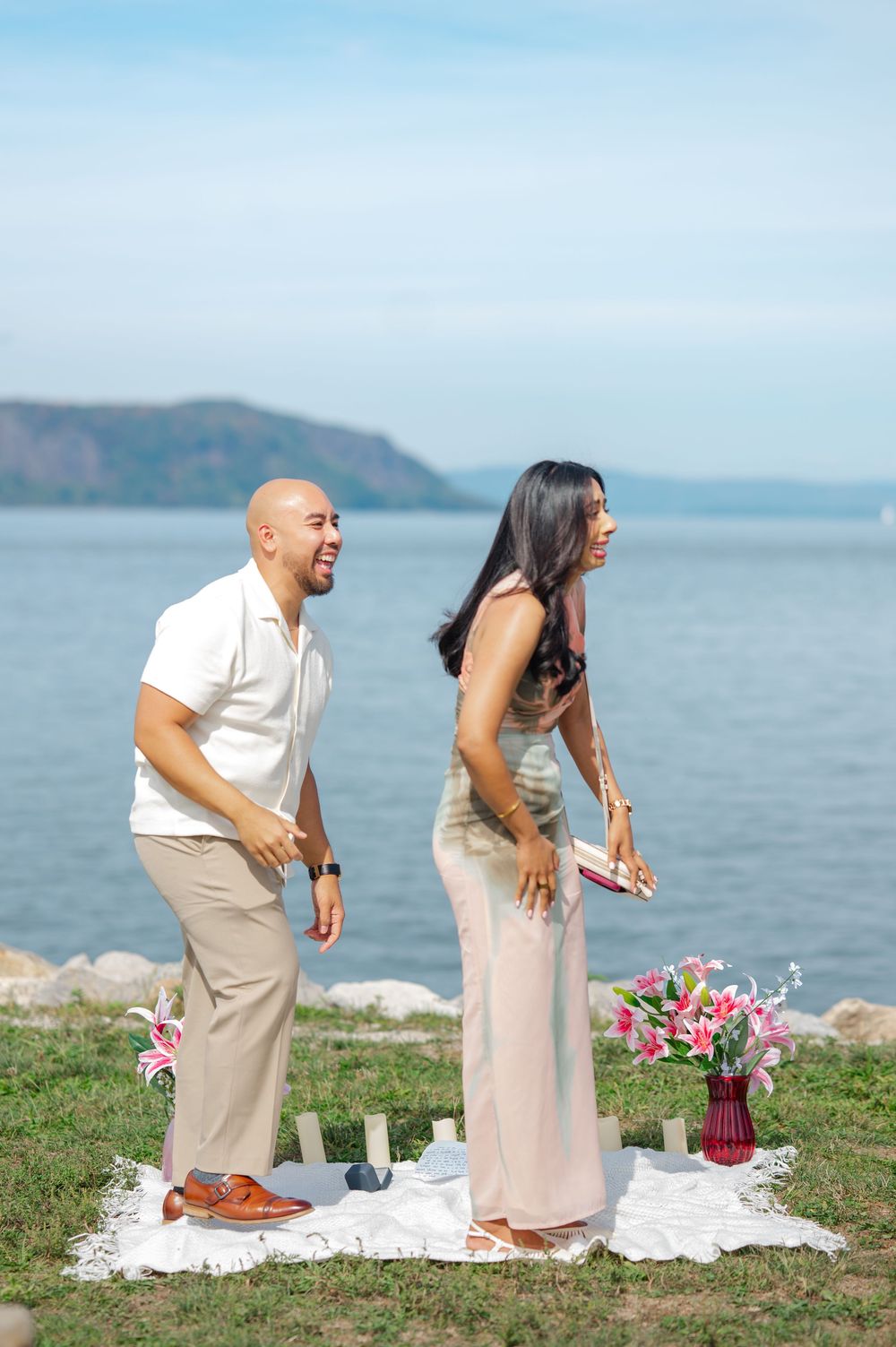 A couple shares a joyful moment during a picnic by the ocean with flowers and a white blanket on the grass.