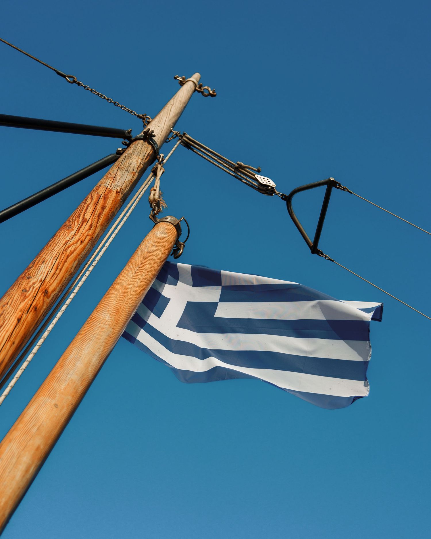A Greek flag flutters against a bright blue sky, mounted on a wooden pole with visible electrical wires nearby.