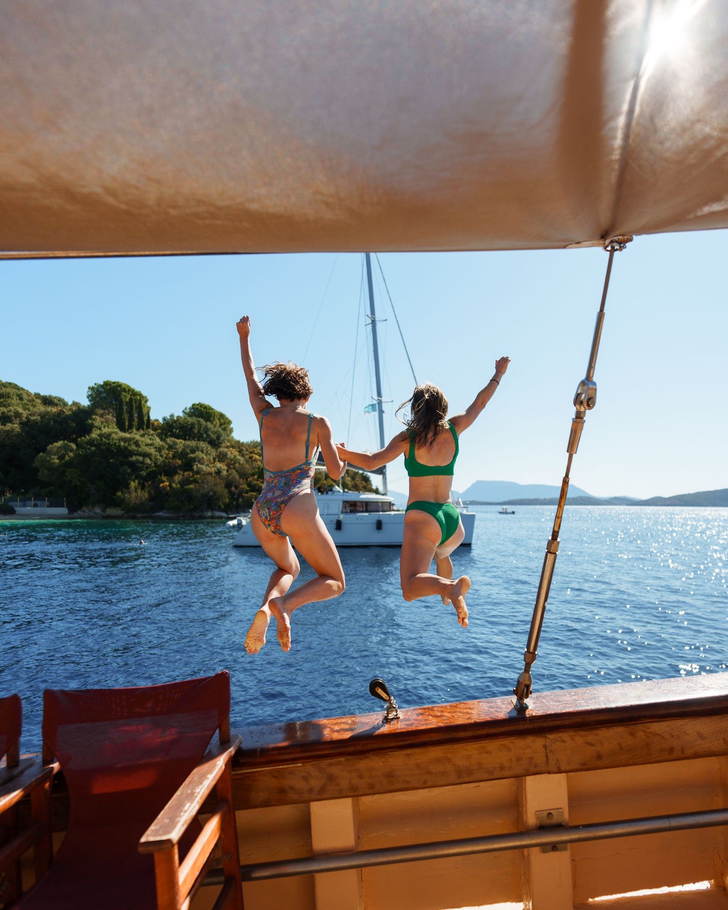 Two people leap off the back of a boat into a scenic lake surrounded by trees on a sunny summer day.