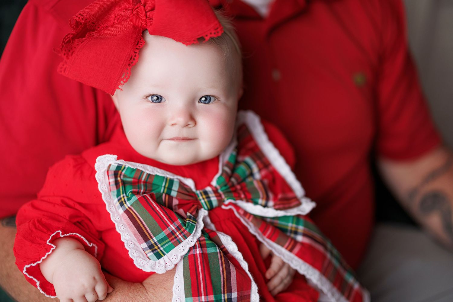 A baby wearing festive holiday attire with a red and green plaid bow sits against a red background.