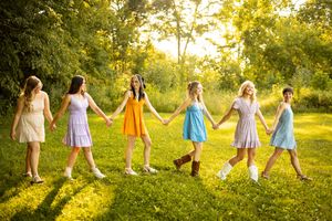 A group in colorful summer dresses holds hands while walking through a sunlit garden at golden hour.