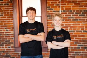 Two people wearing matching black t-shirts stand with arms crossed in front of a brick wall.