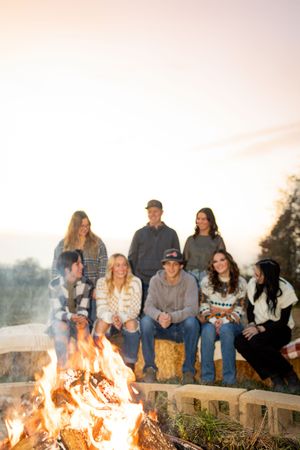 Group of friends gathered around a warm bonfire at sunset, sitting on hay bales in a rustic outdoor setting.