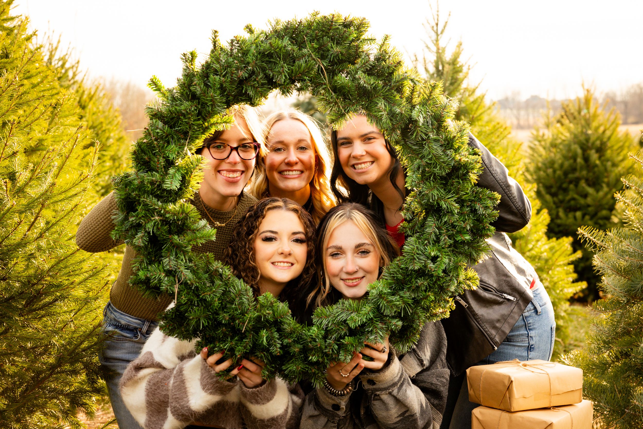 A group poses playfully looking through a large circular holiday wreath at a Christmas tree farm.