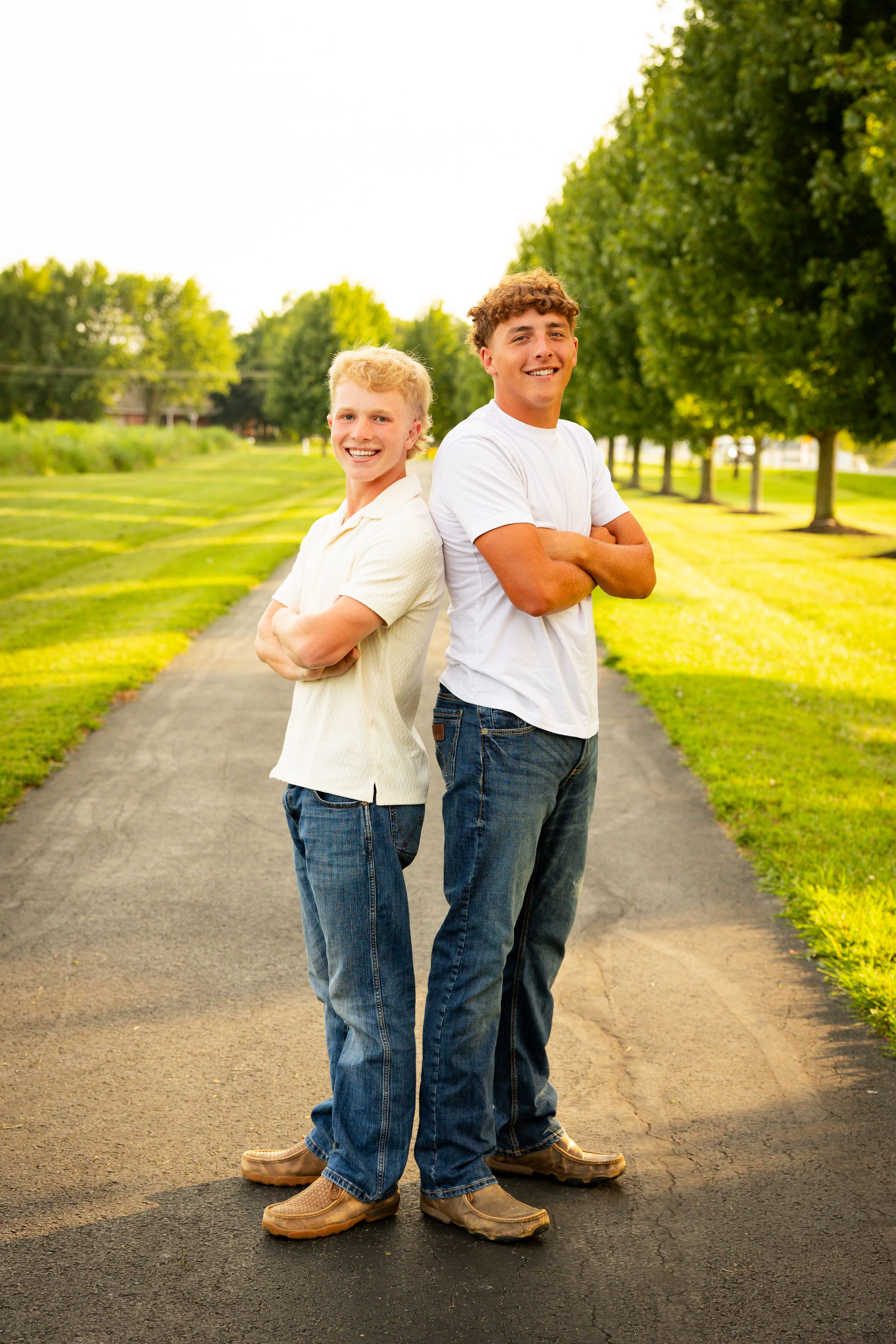 Two young siblings embrace while standing on a path surrounded by green trees and grass in golden sunlight.