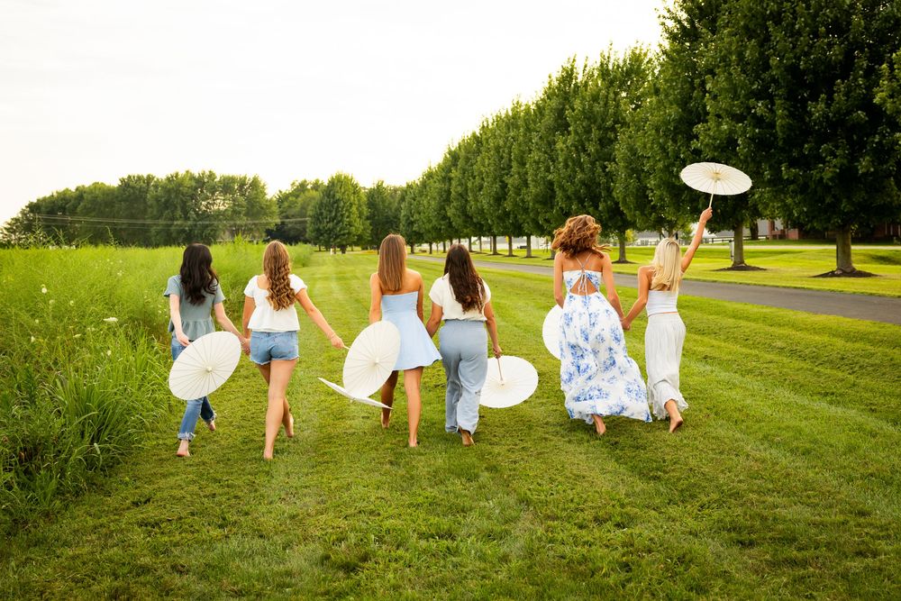 People carrying white parasols walk together across a sunny green field during a summer outdoor gathering.