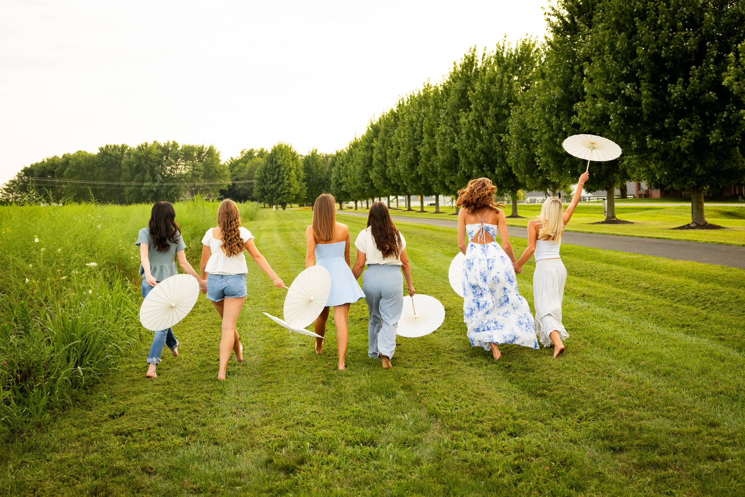 People carrying white parasols walk together across a sunny green field during a summer outdoor gathering.