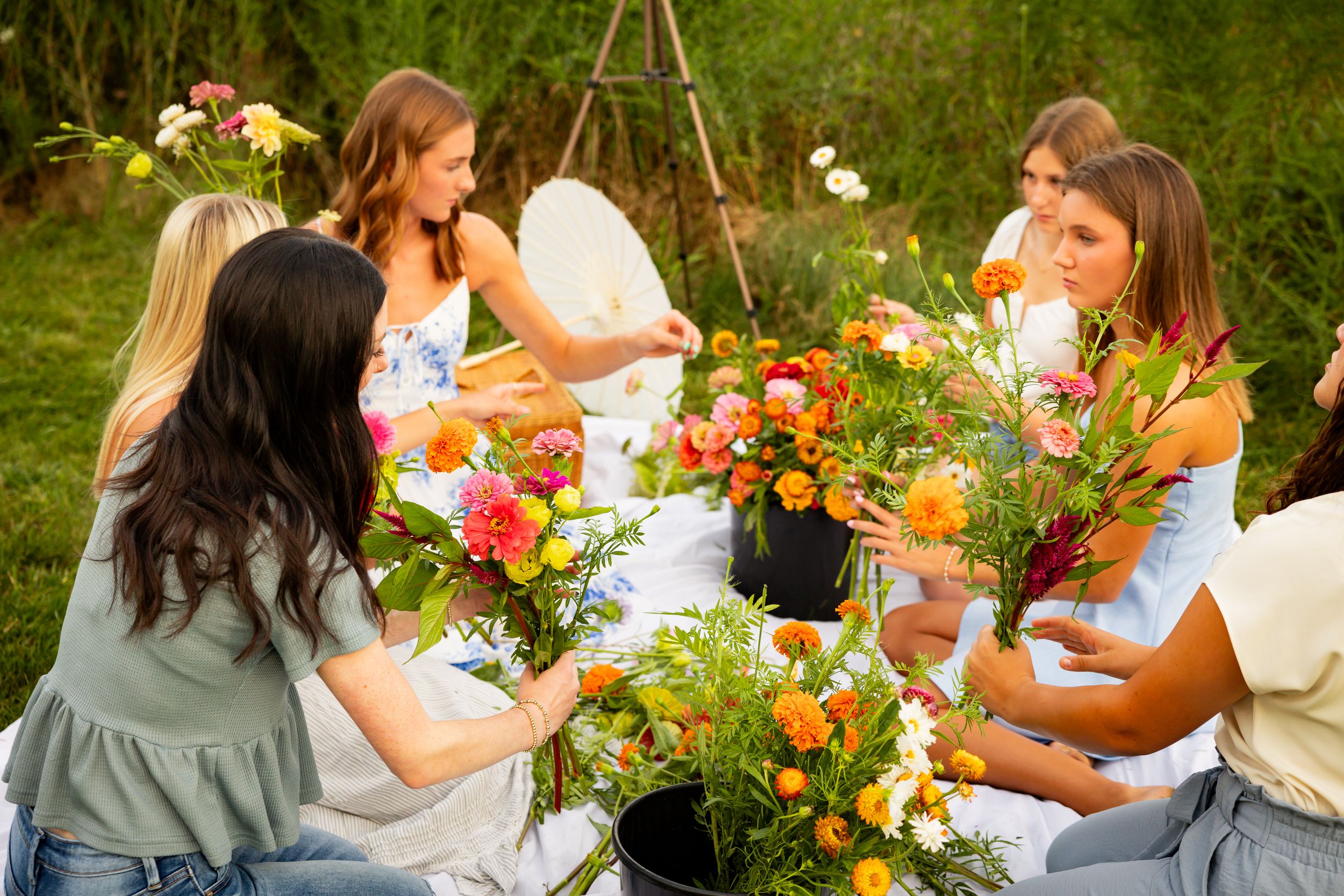 People gather outdoors for a flower arranging workshop with colorful blooms and greenery on a sunny day.