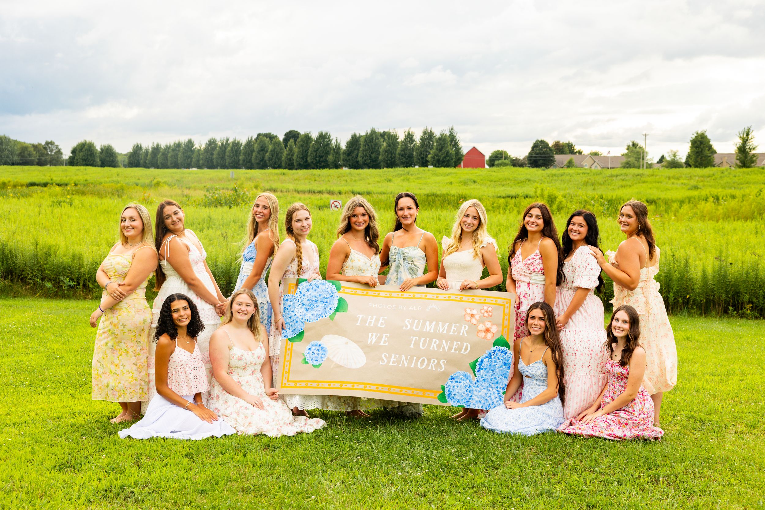 Group of bridesmaids in colorful dresses posing outdoors on green lawn holding decorative sign at wedding celebration.