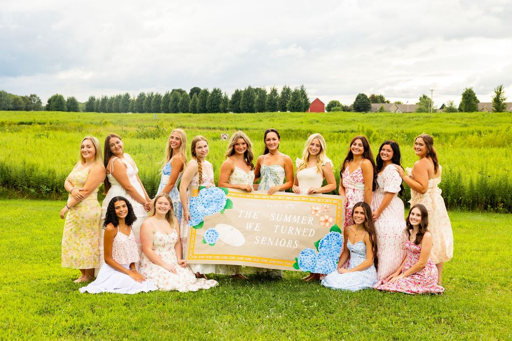 Group of bridesmaids in colorful dresses posing outdoors on green lawn holding decorative sign at wedding celebration.