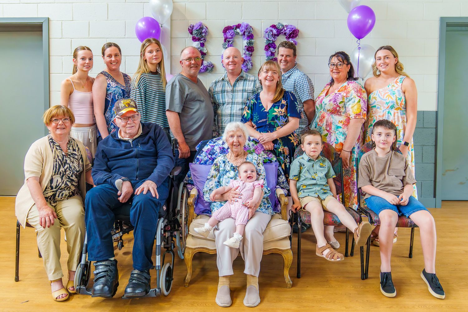 Large multigenerational family gathers to celebrate a 100th birthday with purple balloons and decorations.