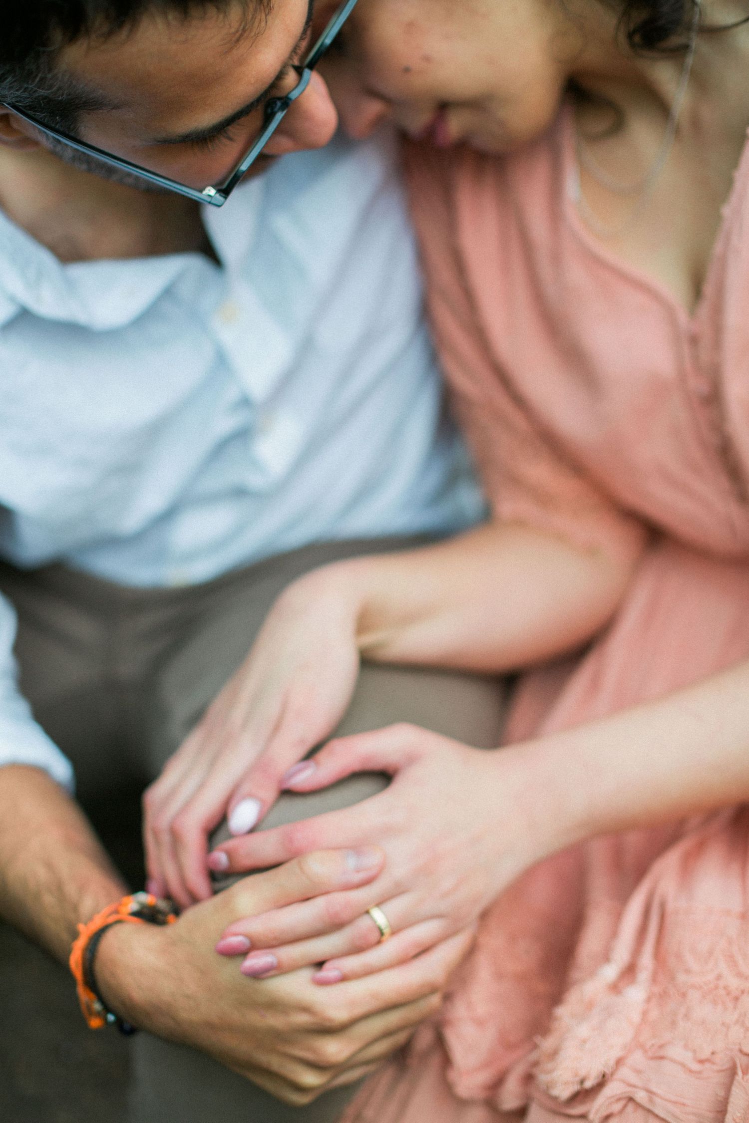Close-up photo is newly engaged couple's hands showing engagement ring on finger with pink dress in background. Hayley Elizabeth Engagements