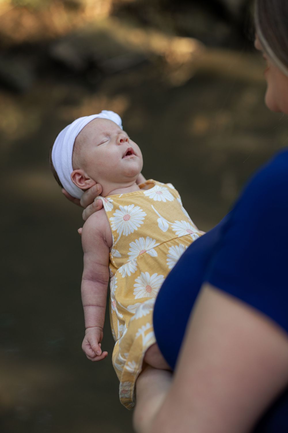 A mother holds her newborn baby girl in a yellow floral dress and white headband outdoors near a creek.