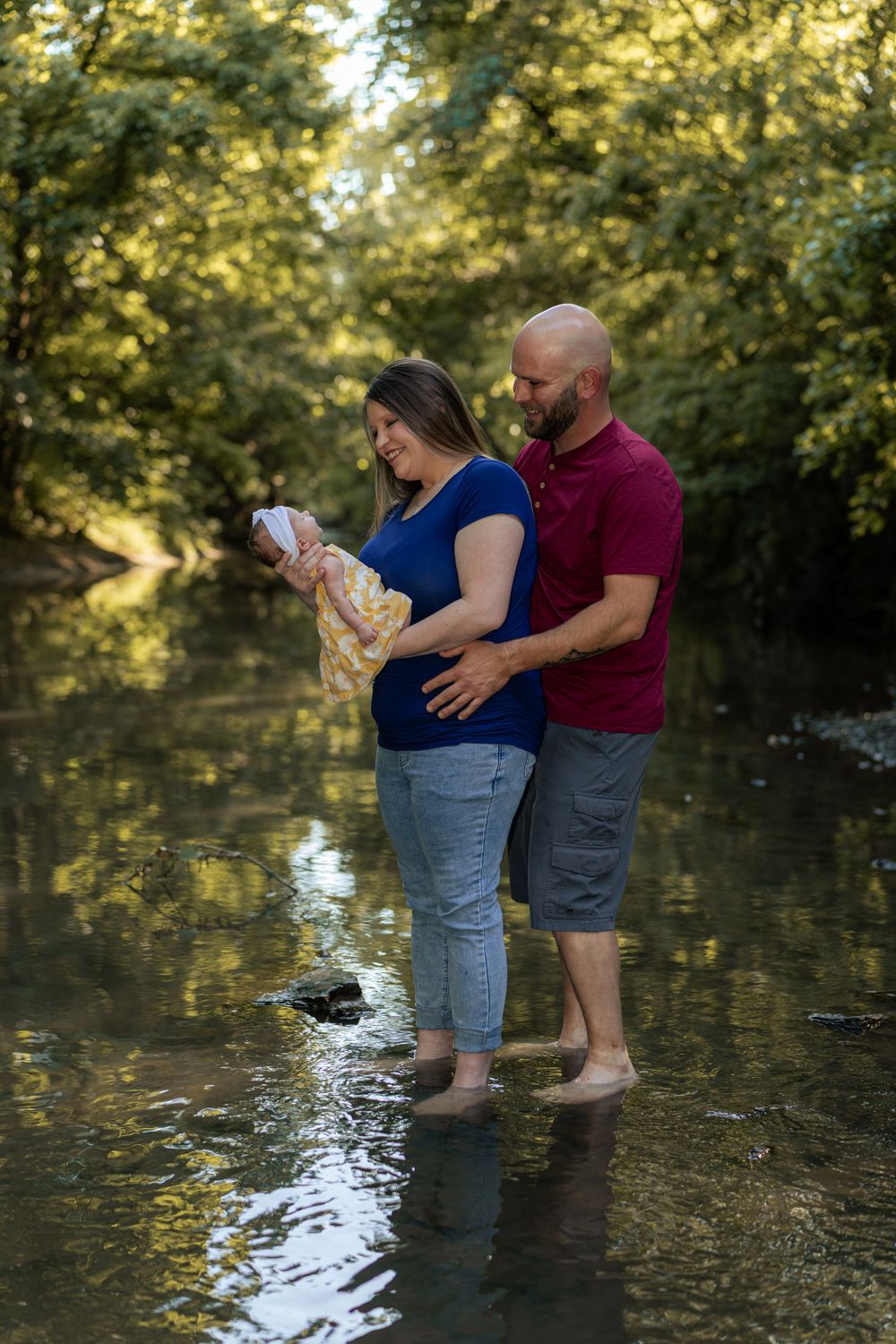 A couple stands barefoot in a shallow creek, lovingly gazing at their newborn baby, surrounded by lush green trees.