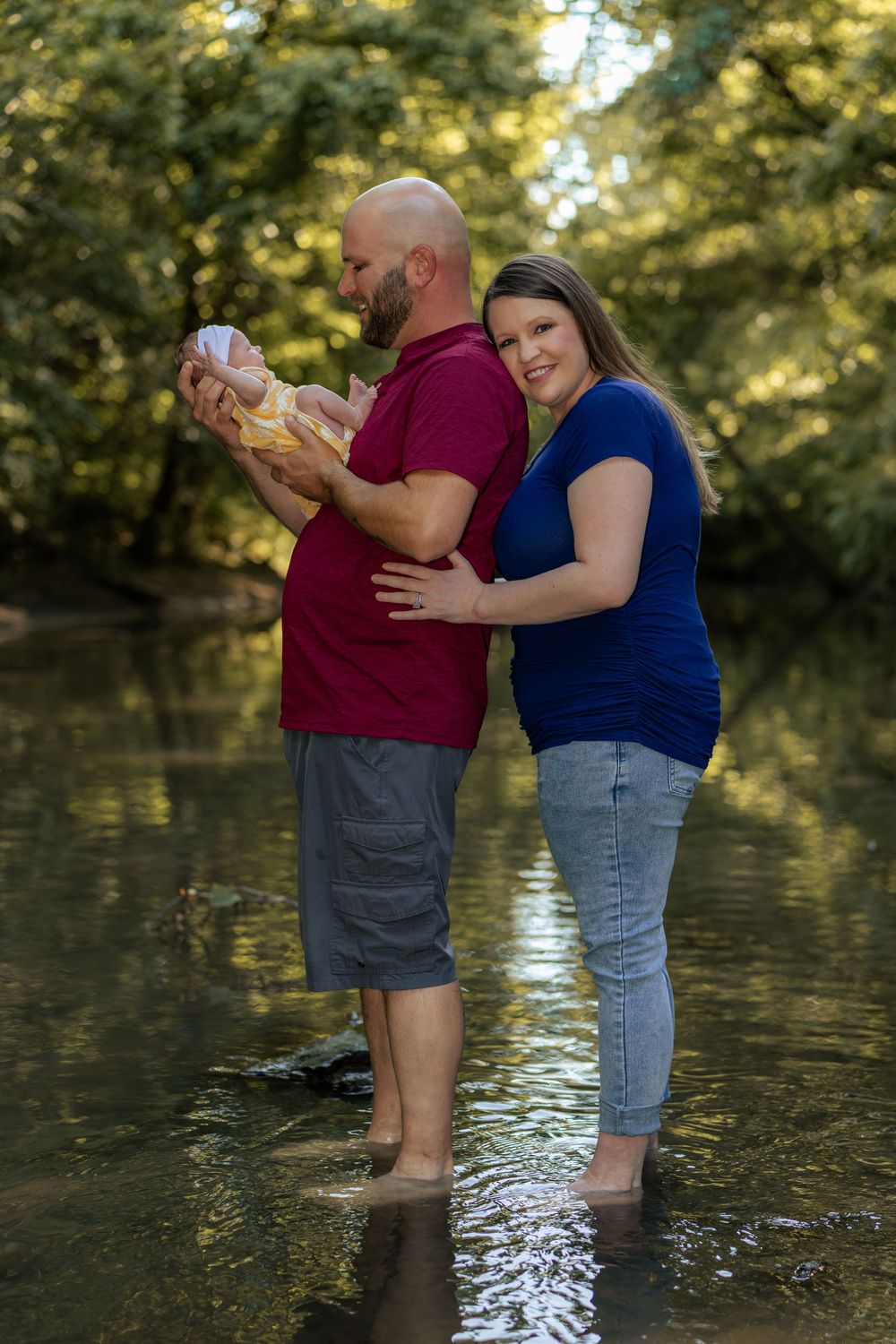 A couple stands barefoot in a shallow creek, with the man holding their newborn baby in a yellow dress.