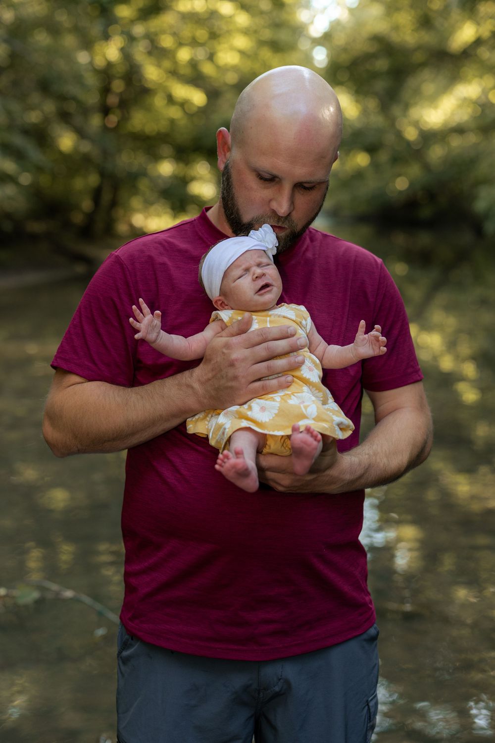 A bald bearded father tenderly kisses his newborn baby in a yellow floral dress and white headband outdoors.