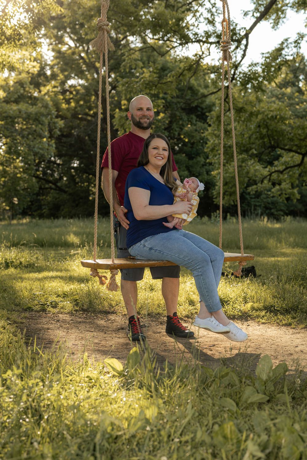 A couple sits on a wooden swing outdoors, the woman holding a newborn baby, surrounded by lush green trees in golden sunlight.