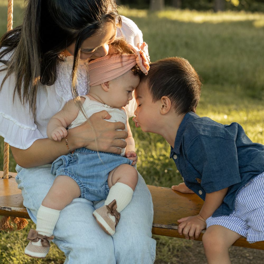 A mother sits on a wooden swing holding her baby girl while a toddler boy leans in for a sweet nose kiss outdoors.