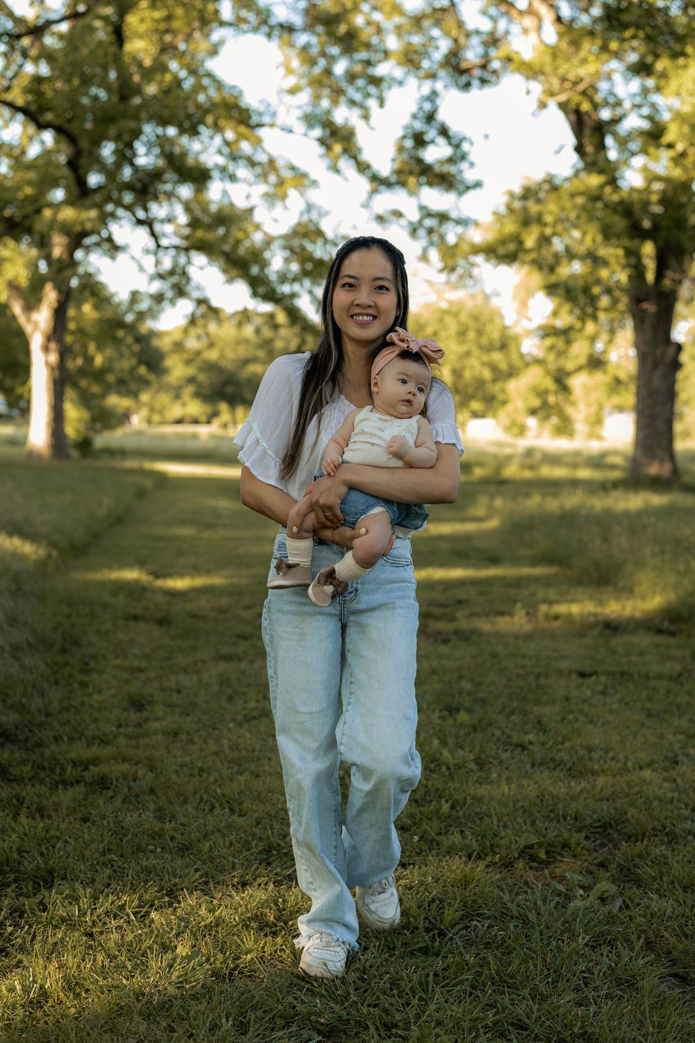 Smiling young woman holding a baby girl while walking in a sunny green park with large trees.
